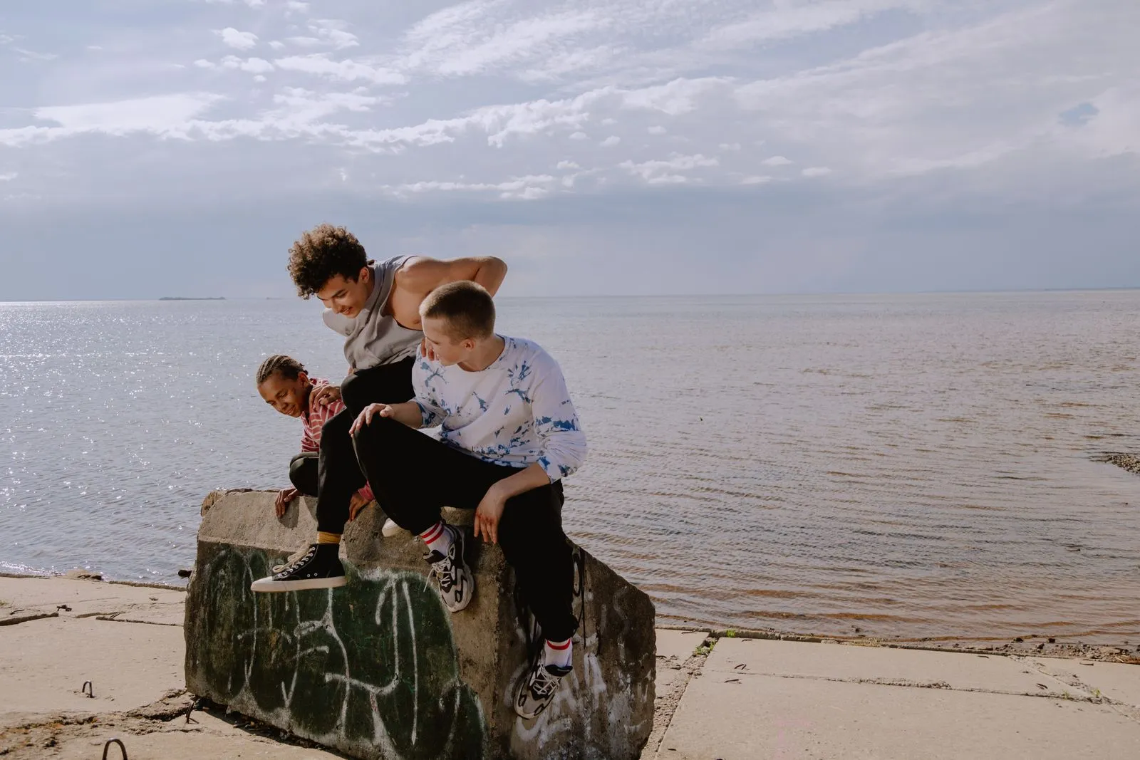 Three diverse teens enjoy a casual day by the seaside, sitting on a concrete block.